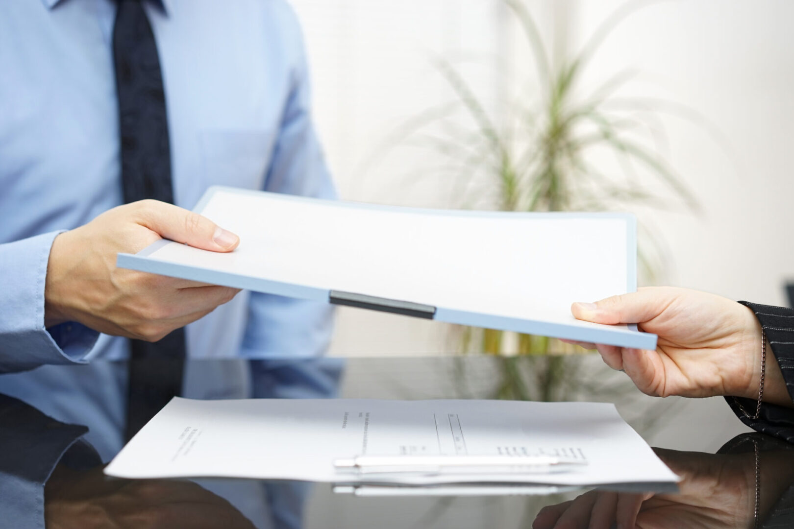 Two individuals are exchanging a clipboard with documents over a desk, possibly discussing court reporting services. A lush plant adds a touch of greenery in the background, creating an atmosphere of professionalism and calm.