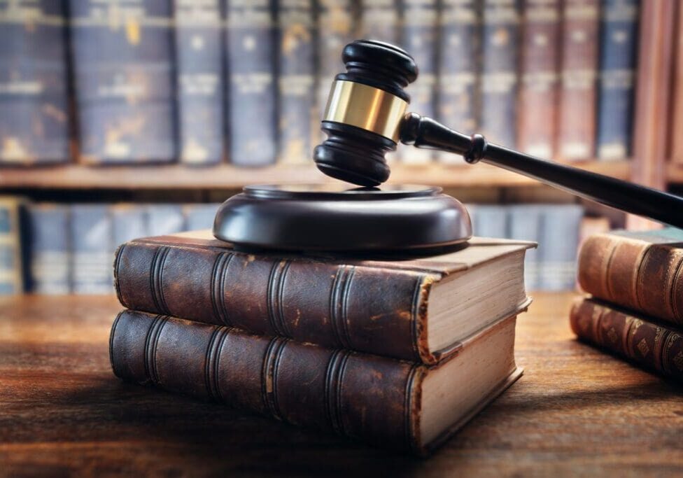 A judge's gavel rests on a sound block atop a stack of three old law books, with blurred bookshelves in the background.
